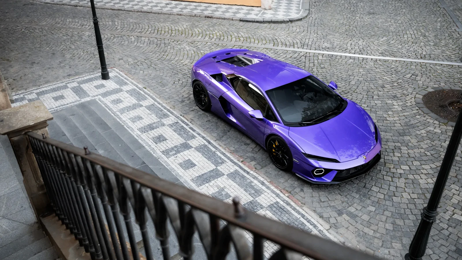 A purple Lamborghini Temerario seen from above on a cobbled Prague street, beside old stone stairs and a street lamp